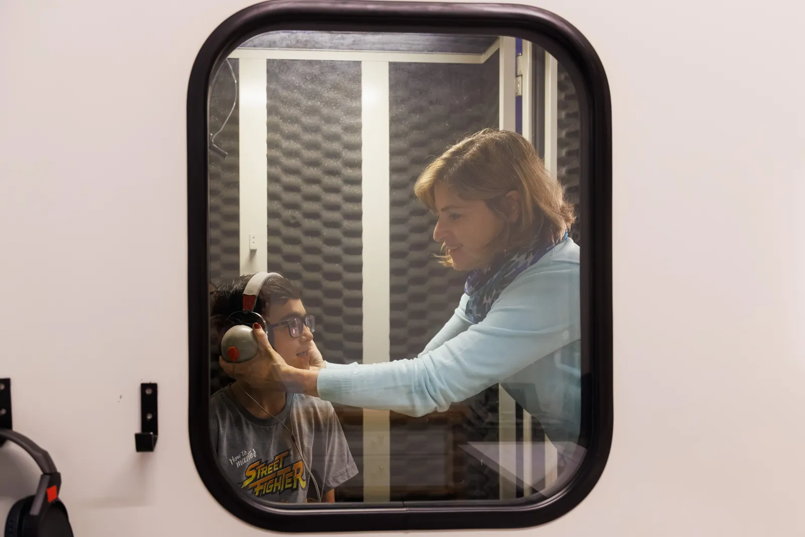 Simone ajustando o fone no paciente dentro da cabine audiométrica, visto pela janela